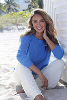 Woman in blue jumper and white trousers sitting on beach