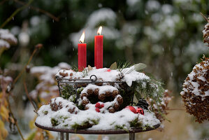 Verschneites Adventsgesteck im Metallkorb mit roten Kerzen und Tannenzapfen