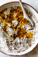 An overhead view of a ceramic bowl filled with a mixture of flour, seeds, and liquid ingredients, with a wooden spoon resting inside.