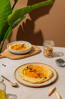 A rustic table setting with creamy hummus drizzled with olive oil and sprinkled with seeds. Bright sunlight emphasises the texture and decorative leaves lie nearby.