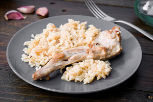 A tasty dish of fried rabbit served with creamy rice on a grey plate Rustic wooden table decoration with garlic cloves and a fork to enhance the presentation