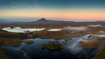 Luftaufnahme eines ruhigen Sees, in dem sich der Sonnenuntergang mit majestätischen Bergen und einem heiteren Himmel spiegelt, Glencoaghan, Bencorr, County Galway, Irland.