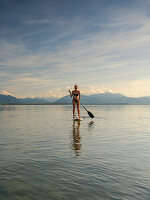 A young woman doing stand-up paddling on the Chiemsee, in the background the Chiemgau Alps, Chieming, Upper Bavaria, Germany