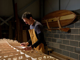 Man at work standing over wooden paddleboard in workshop holding paper and pencil