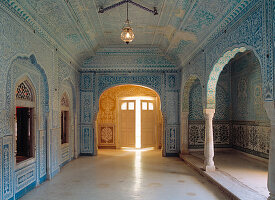 A striking blue room, detailed with lavish wall decorations, looking towards a door, opening into the outside and a blazing sunlight