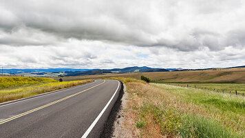 A blacktop road through a flat landscape with undulating hills, farmland and rolling hills beyond.