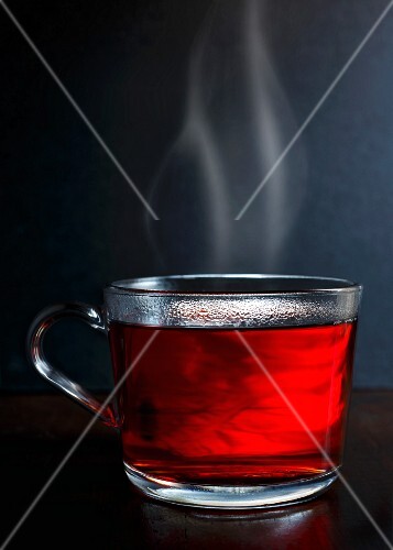 Hot red berry fruit tea in a glass mug with steam standing on a dark wood with dark grey background